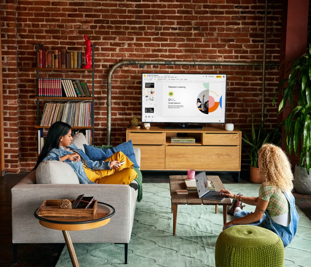 A person sits on a sofa watching a TV presentation. A second person sits on the floor at a coffee table with a Chromebook.