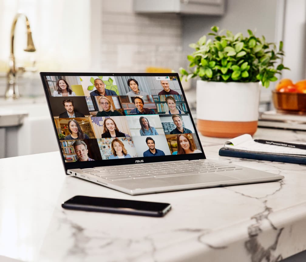 A Chromebook sits open on a white marble worktop with a mobile phone on the left and a plant in the background.